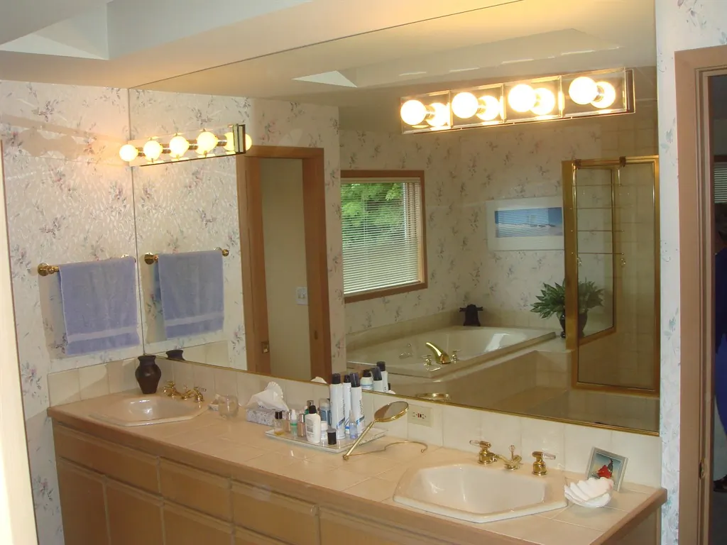 Bathroom before remodel showing tile vanity countertops, large wall mirror, floral wallpaper, built-in tub, and gold-framed shower enclosure